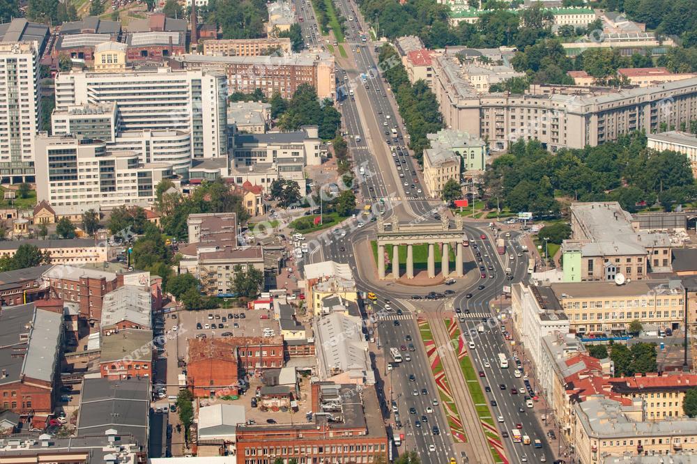 На фотографии изображены Московские триумфальные ворота, расположенные в Санкт-Петербурге, в обрамлении современных и исторических зданий. Панорамный вид с высоты демонстрирует структуру городской застройки и оживлённые магистрали. Цветущие газоны и зелень деревьев подчёркивают летнюю атмосферу. Этот архитектурный памятник символизирует историческую связь города с важными событиями прошлого.